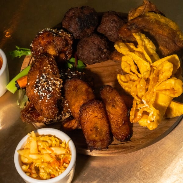 A wooden platter with Caribbean appetizers: chicken wings, fried plantains, fritters, and wontons.